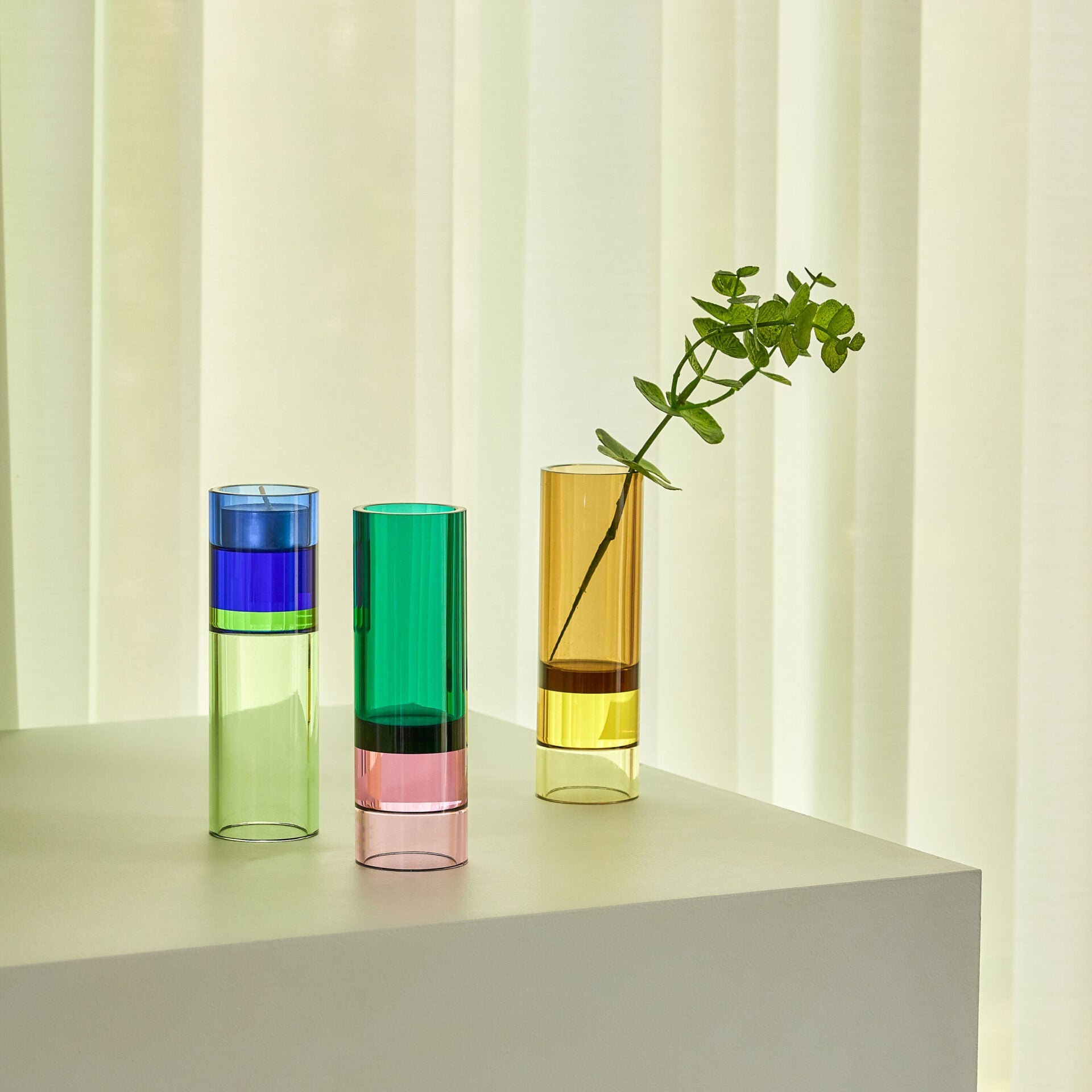 drei zylindrische glas-teelichthalter in grün, blau und bernsteinfarben, auf einem hellen tisch vor hellen vorhängen, einer mit grünem zweig als vase verwendet
