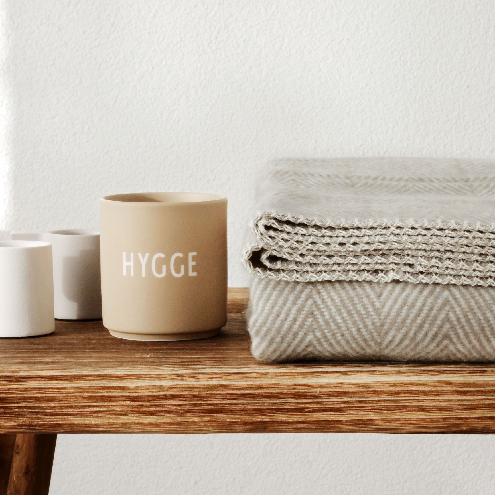 Gefaltete taupefarbene Baumwolldecke mit Fischgrätmuster neben Tasse mit „HYGGE“-Aufdruck auf Holztisch