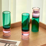 drei hohe zylindrische glas-teelichthalter in grün und rosa auf einem holztablett vor hellen vorhängen, einer mit brennendem teelicht