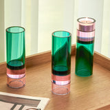 drei hohe zylindrische glas-teelichthalter in grün und rosa auf einem holztablett vor hellen vorhängen, einer mit brennendem teelicht