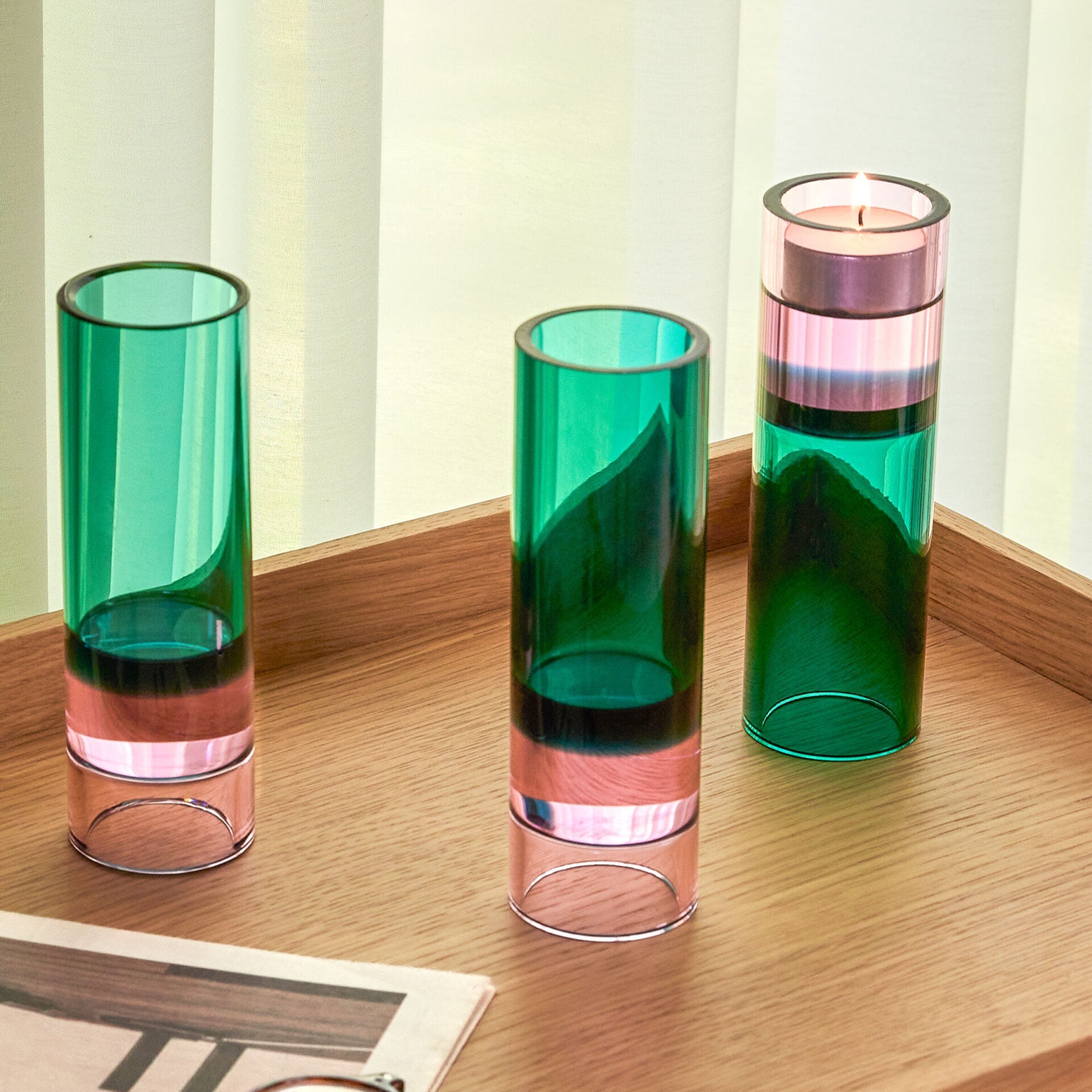 drei hohe zylindrische glas-teelichthalter in grün und rosa auf einem holztablett vor hellen vorhängen, einer mit brennendem teelicht