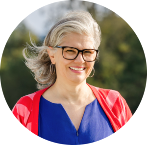 portraitfoto von claudia mit grauen haaren, brille und blauem oberteil mit roter jacke draußen im grünen