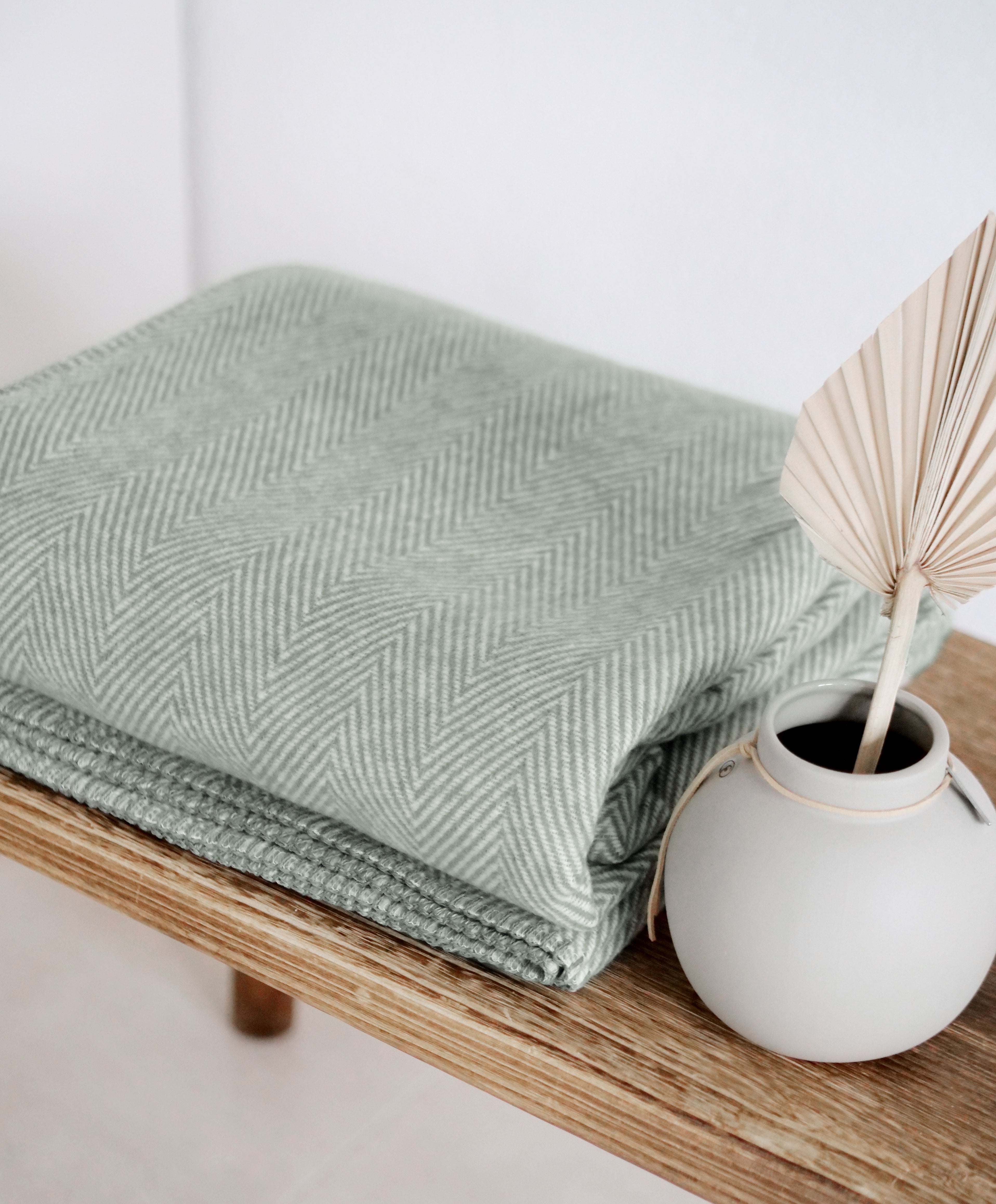 Grüne Riema Germany Kuscheldecke aus Biobaumwolle im Fischgrätmmuster auf Holzbank mit Vase

