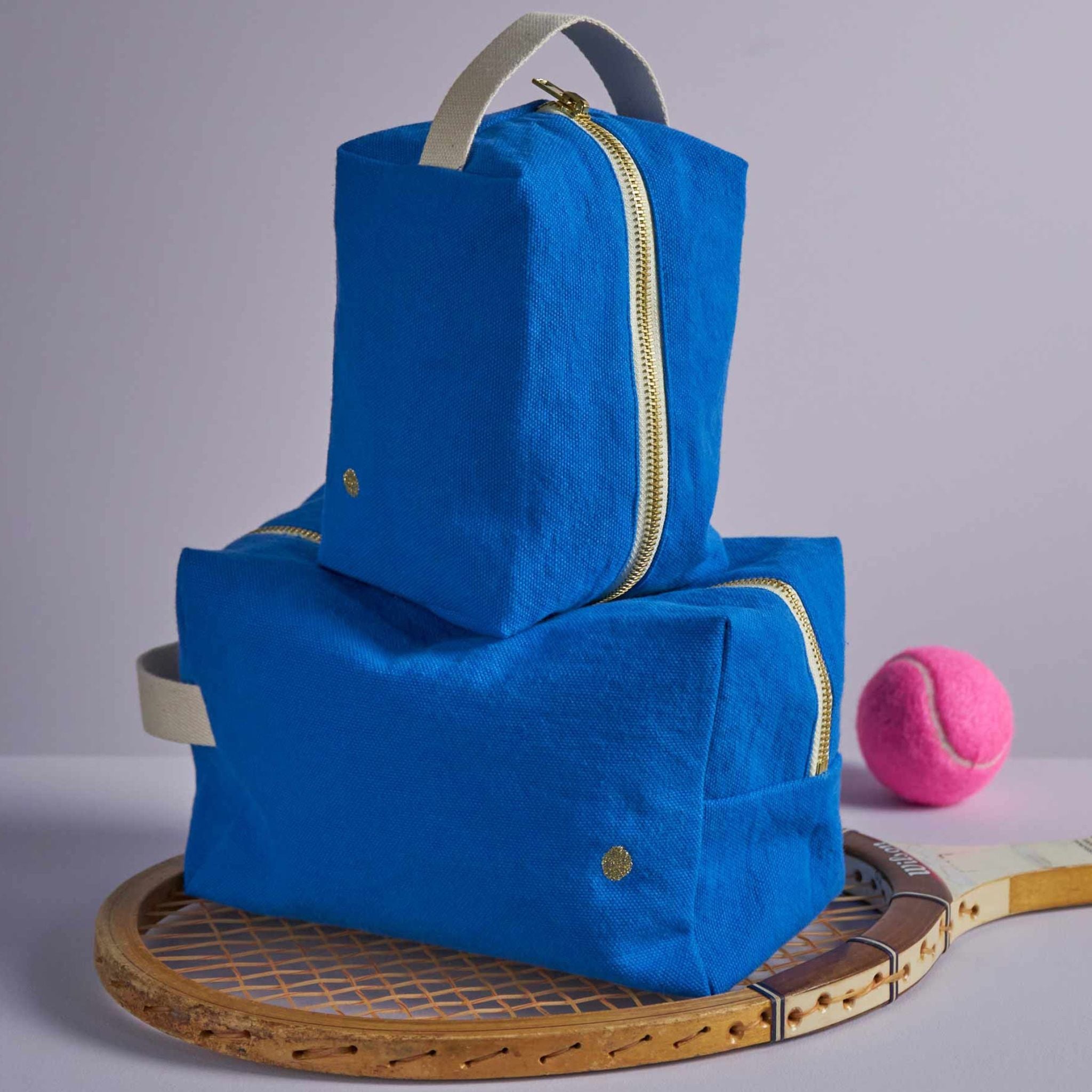 Zwei Kulturbeutel in Würfelform in blau aus Bio-Baumwolle mit Pflegeprodukten befüllt auf einem Tennisschläger fotografiert.
